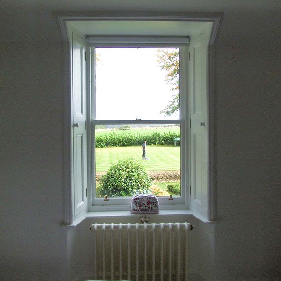 Inside of room looking towards traditional window with wooden shutters and garden scene through the window panes